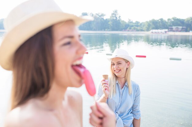 Free photo woman licking popsicle in front of her friend holding icecream cone near the lake