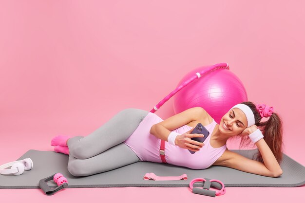 woman lies on comfortable fitness mat watches video via smartphone dressed in sportswear exercises with sport equipment 