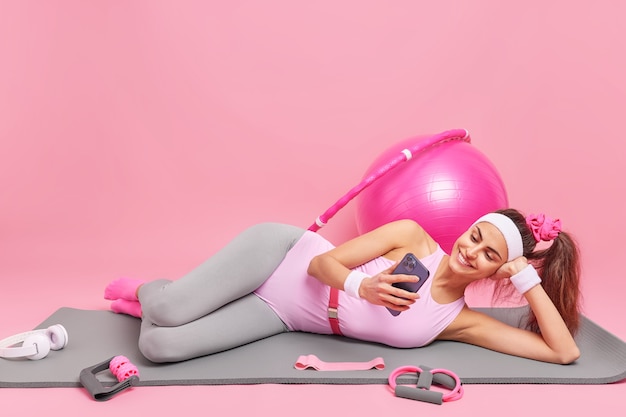 woman lies on comfortable fitness mat watches video via smartphone dressed in sportswear exercises with sport equipment 