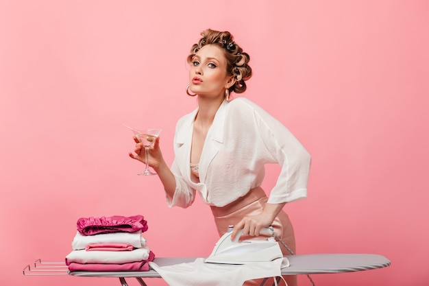 Woman in light silk outfit posing on pink wall with martini glass and ironing clothes