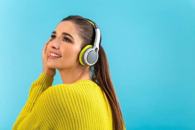 woman listening to music in wireless headphones happy wearing yellow knitted sweater posing on blue