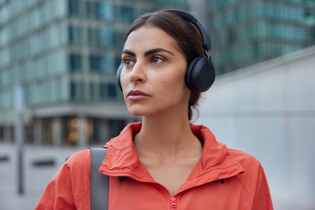 Free photo woman listens music in wireless headphones enjoys audio track dressed in anorak waits for coach poses outdoors