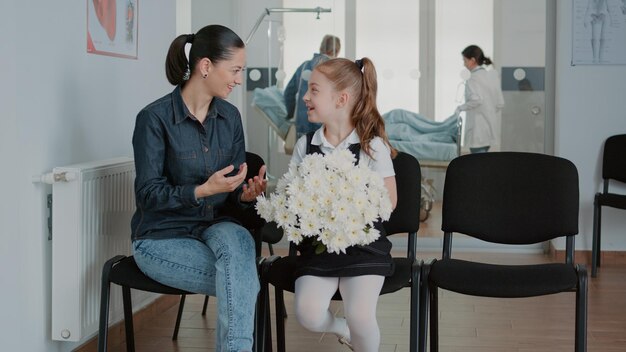 Woman and little girl preparing to visit patient in waiting room, having flowers for sick person. Mother and child waiting to enter hospital ward at medical facility. People in waiting area