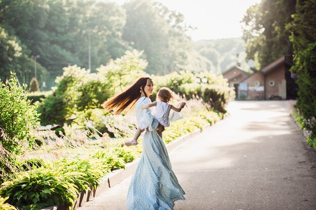 Woman in long blue dress whirls with her little daughter on pavement path 