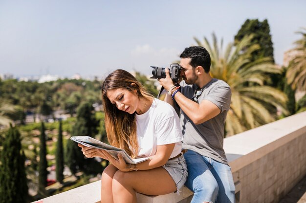 Woman looking at map sitting with her boyfriend taking photograph on camera in the park