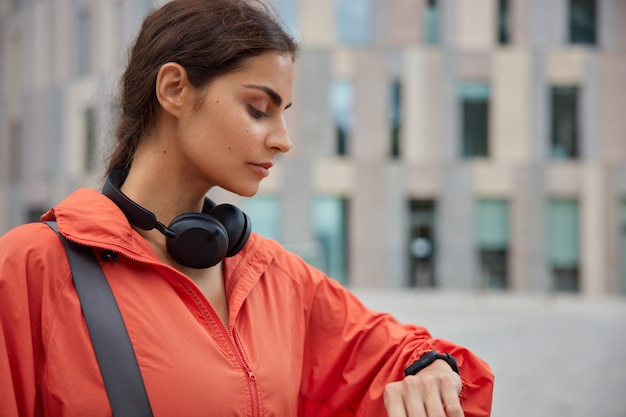 Free photo woman looks at activity fitness tracker has workout break running application on smartwatch monitors her sport activity stands outdoor on blurred city view.