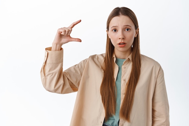 Free Photo woman looks confused while shows tiny small gesture, little thing on copyspace, stare at front puzzled, disappointed with size, standing over white wall