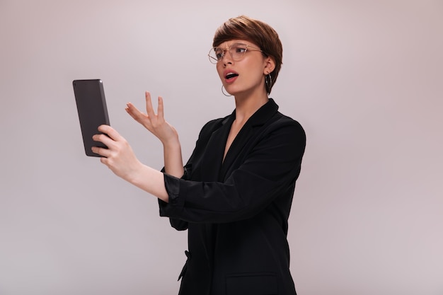 Woman looks into tablet screen with misunderstanding. Business lady in black jacket poses on isolated white background