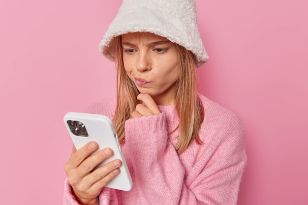 woman looks at smartphone screen reads notification receives message from unknown person wears white hat and jumper isolated on pink has doubtful expression