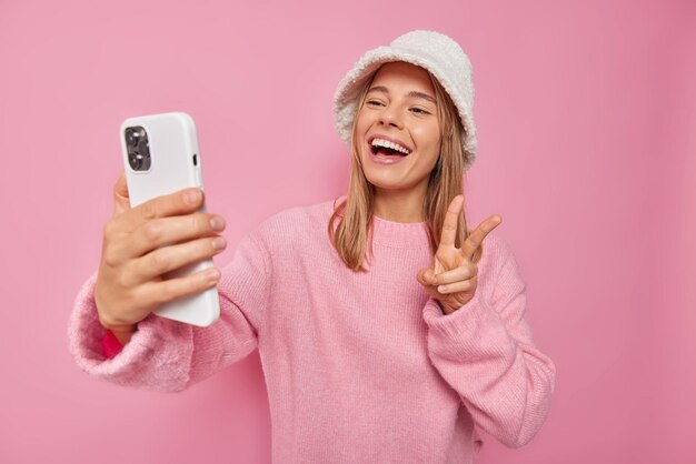 woman makes self portrait via cellphone shows peace sign smiles gladfully wears casual jumper and hat poses on pink