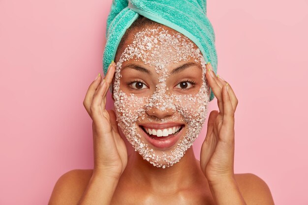 Woman massages face, applies natural scrub mask, cleans facial skin for having young look, has manicure, wears wrapped towel on head, isolated on pink wall