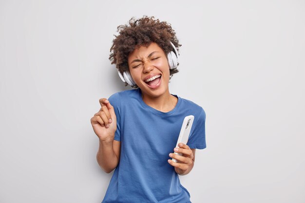 woman meloman enjoys favorite music via headphones holds mobile phone has fun moves with rhythm of favorite music wears casual blue t shirt isolated on white