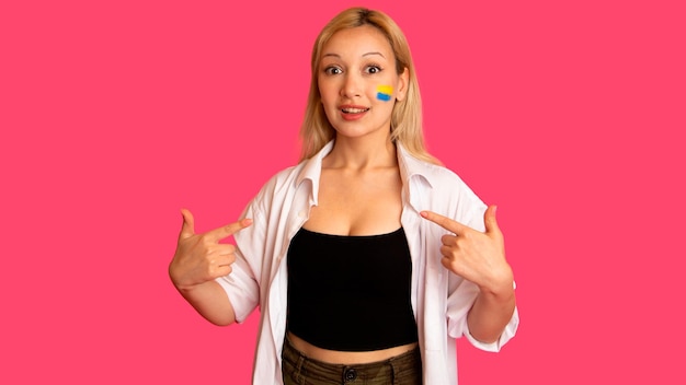 Woman of model appearance with the flag of Ukraine on her face holds posing on a pink background