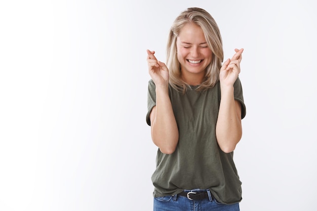 woman in olive t-shirt close eyes smiling thrilled and amused holding fingers crossed for good luck as making wish aganst white