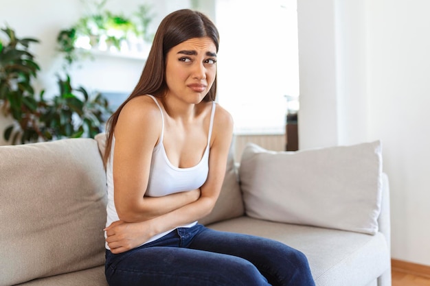 Woman in painful expression holding hands against belly suffering menstrual period pain lying sad on home bed having tummy cramp in female health concept