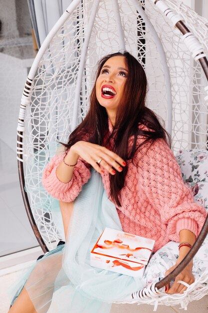 Woman in pink sweater and blue skirt stand by egg chair holding candy box