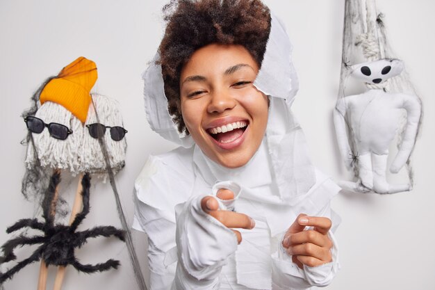  woman points directly at camera collects accessories for carnival enjoys October time and spooky holiday wears ghost costume smiles broadly poses indoor on white 