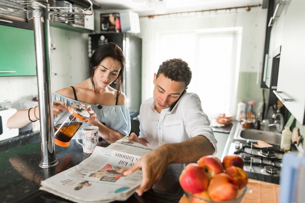 Woman pouring coffee in mug sitting near the husband talking on mobile phone