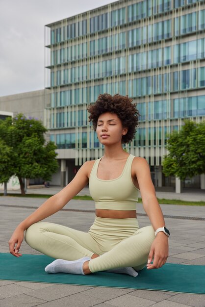 woman practices yoga lesson poses with mudra gesture on rubber mat dressed in tracksuit being in urban place