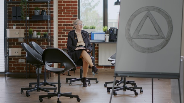 Woman psychiatrist waiting on people to attend aa group therapy meeting. Psychotherapist sitting in circle with chairs and holding clipboard to give advice to rehabilitation patients.