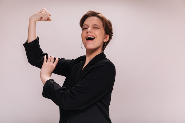 Free photo woman rejoices to her power and demonstrates her biceps. charming lady in black jacket smiles and shows muscles on isolated background