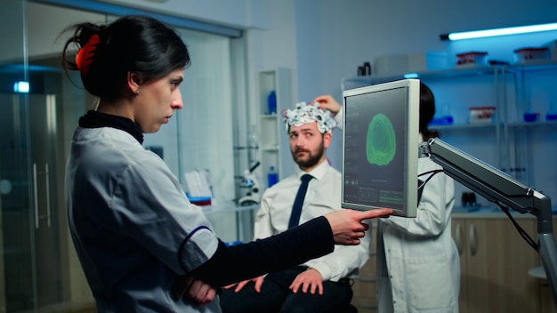 Woman researcher looking at monitor analysing brain scan while coworker discussing with patient in background about side effects