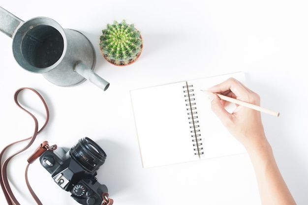 Woman's hand holding pencil on empty notebook with film camera, pot of cactus and vintage watering,  isolated on white background, top view, flat lay