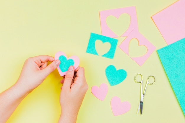 Free photo woman's hand making heart shape with blue and pink paper on yellow background