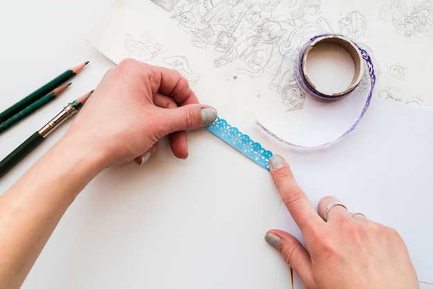 Free photo woman's hand sticking the blue lace on drawing paper over the white backdrop