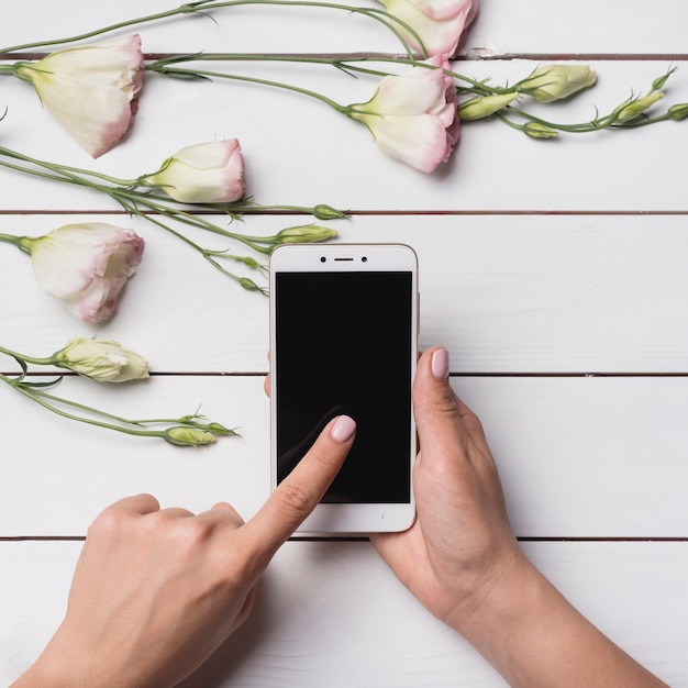 Free photo woman's hand touching the mobile display screen and eustoma flowers over wooden desk