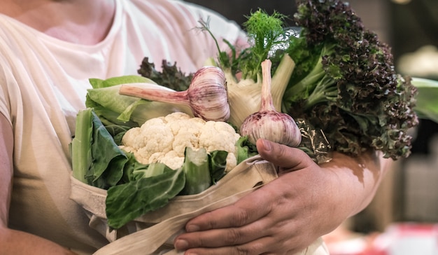 Woman's hands holding fresh ripe organic broccoli, salad with greens and vegetables in cotton bag at the weekend farmer's market