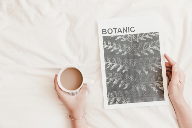 Woman's hands holding hot drink and poster on white bedsheet