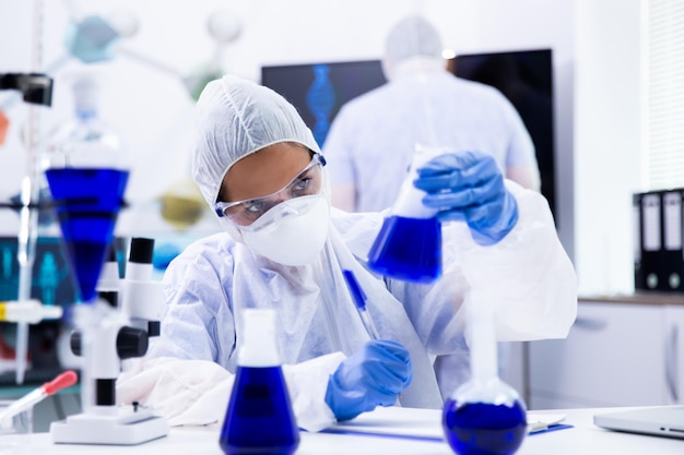 Woman scientist is looking at the beaker with a blue substance inside. Lab colleagues working in the back