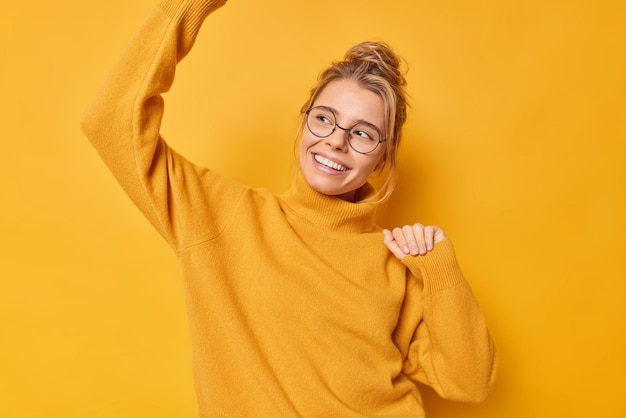 Free photo woman shakes arms dances and enjoys life smiles gently wears spectacles and casual jumper moves with rhythm of music isolated on yellow. monochrome shot