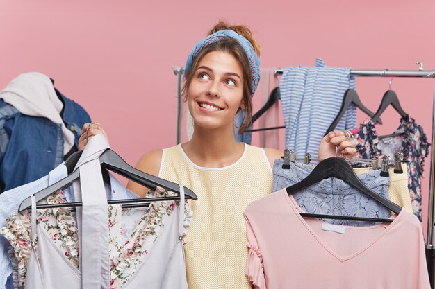 Woman shopaholic being in boutique choosing many outfits, looking with dreamy expression up, not knowing what clothes to choose for date with boyfriend. Joyful female buyer of fashionable clothes