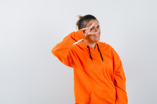 Woman showing V-sign on eye in orange hoodie and looking merry