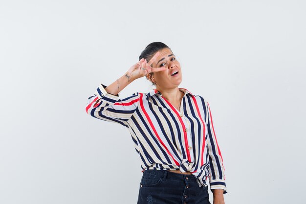 woman showing V-sign near eye in shirt, skirt and looking confident