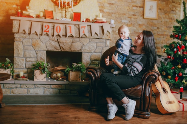 Free Photo woman sitting in an armchair with her baby and a guitar beside and a chimney background