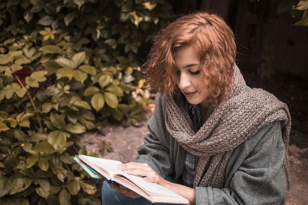 Free photo woman sitting on board and attentively reading book near plant