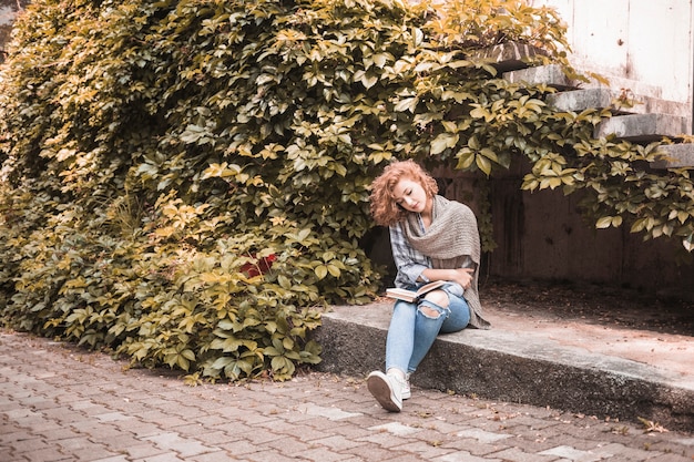 Free photo woman sitting on board and reading book near plant