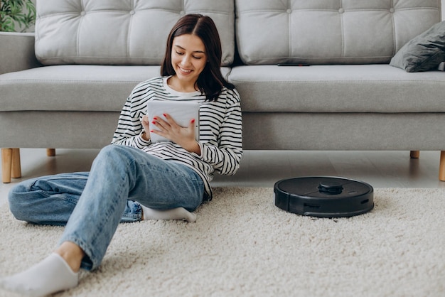 Free photo woman sitting by the sofa using tablet while robot vacuum cleaner cleaning up the room