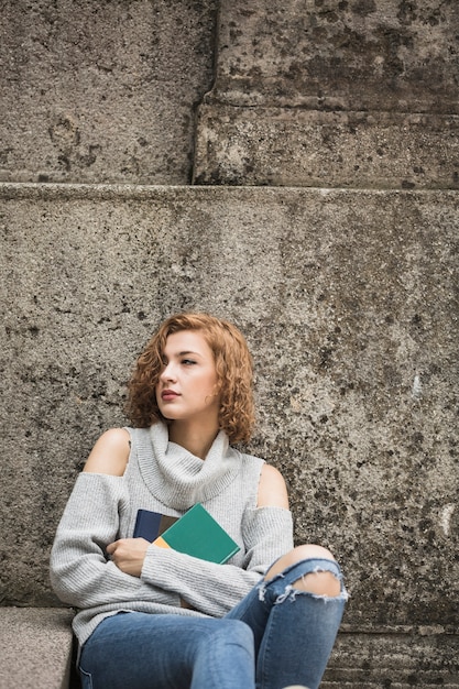 Free photo woman sitting near stone wall and holding books