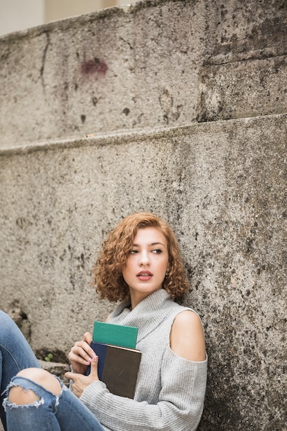 Free photo woman sitting near stone wall and holding notebooks