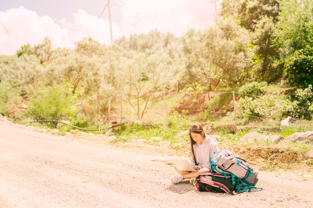 Free photo woman sitting on road and typing on notebook among backpacks