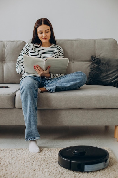 Free photo woman sitting on sofa reading a book while robot vacuum cleaner cleaning up the room