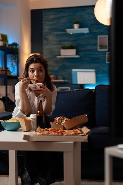 Free Photo woman sitting on sofa watching television while eating takeout asian noodles with chopsticks in home living room. office worker enjoying delicious tv dinner fast food delivery box.