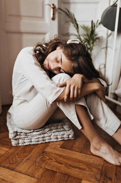 Woman sitting on wooden floor and sleeping Pretty young girl in beige shirt and pants resting in cozy white apartment