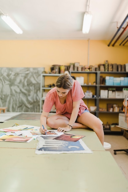 Free photo woman sitting on workbench doing painting on canvas paper in workshop