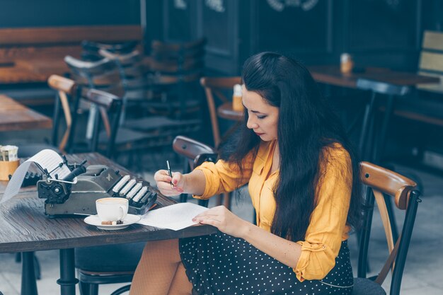 woman sitting and writing something on paper in cafe terrace in yellow top and long skirt during daytime and looking thoughtful