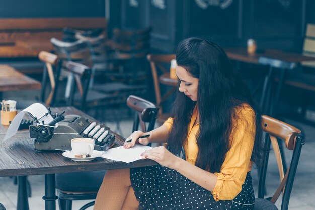 woman sitting and writing something on paper and looking thoughtful and in yellow top and long skirt at cafe terrace during daytime.